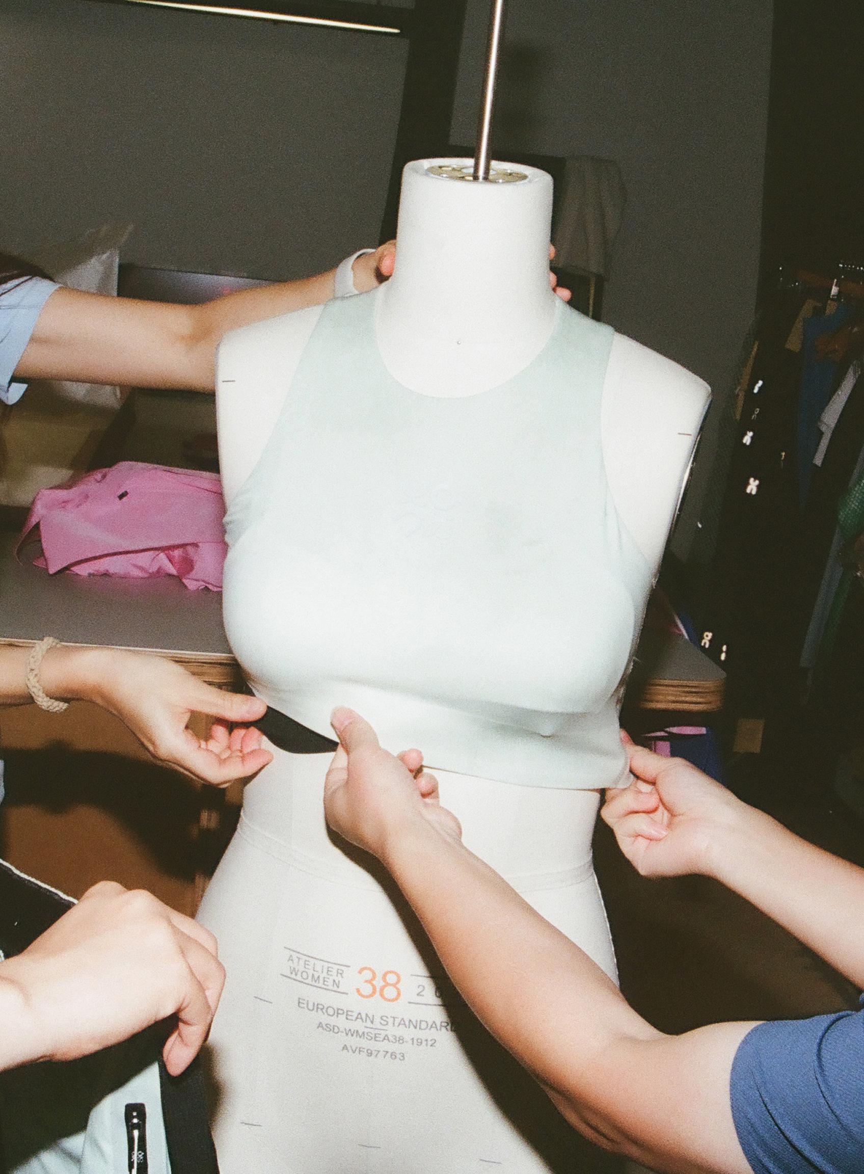 A group of people adjust a mint green crop top on a mannequin while collaborating on a garment fitting.