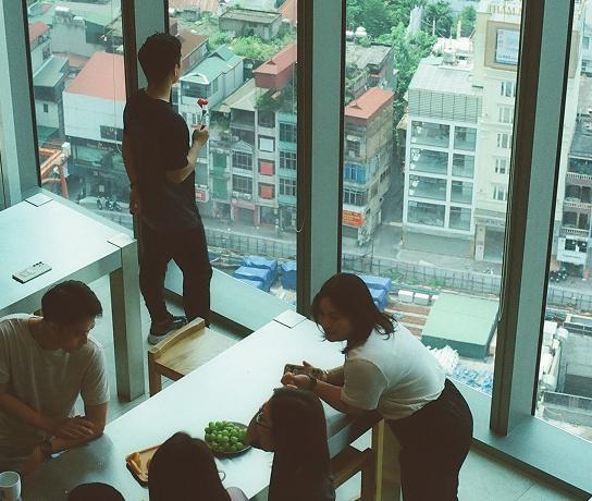 Several people sit and stand around tables in a high-rise room with floor-to-ceiling windows overlooking a city street, one person standing by the window holding a red object on a long stem and a bowl of green fruit on the table.