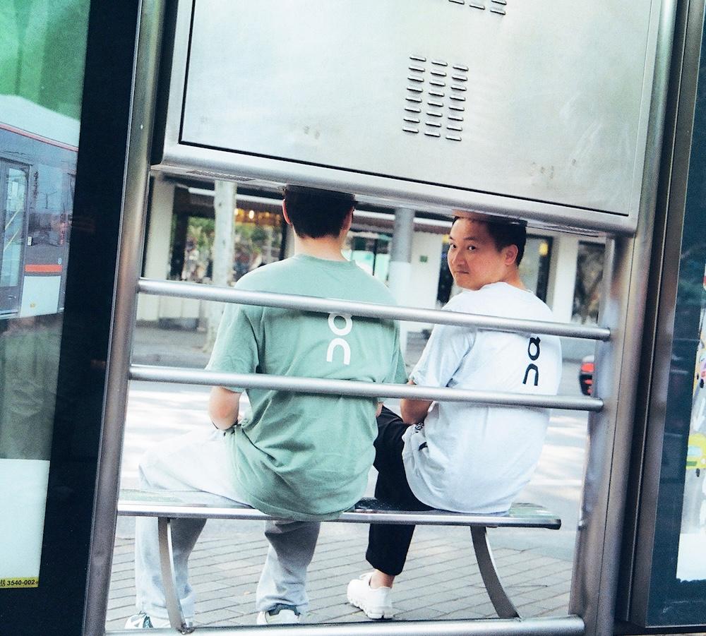 Two people sit side by side on a bench inside a metal-framed shelter, one wearing a green shirt facing away and the other wearing a white shirt turned to look toward the camera.