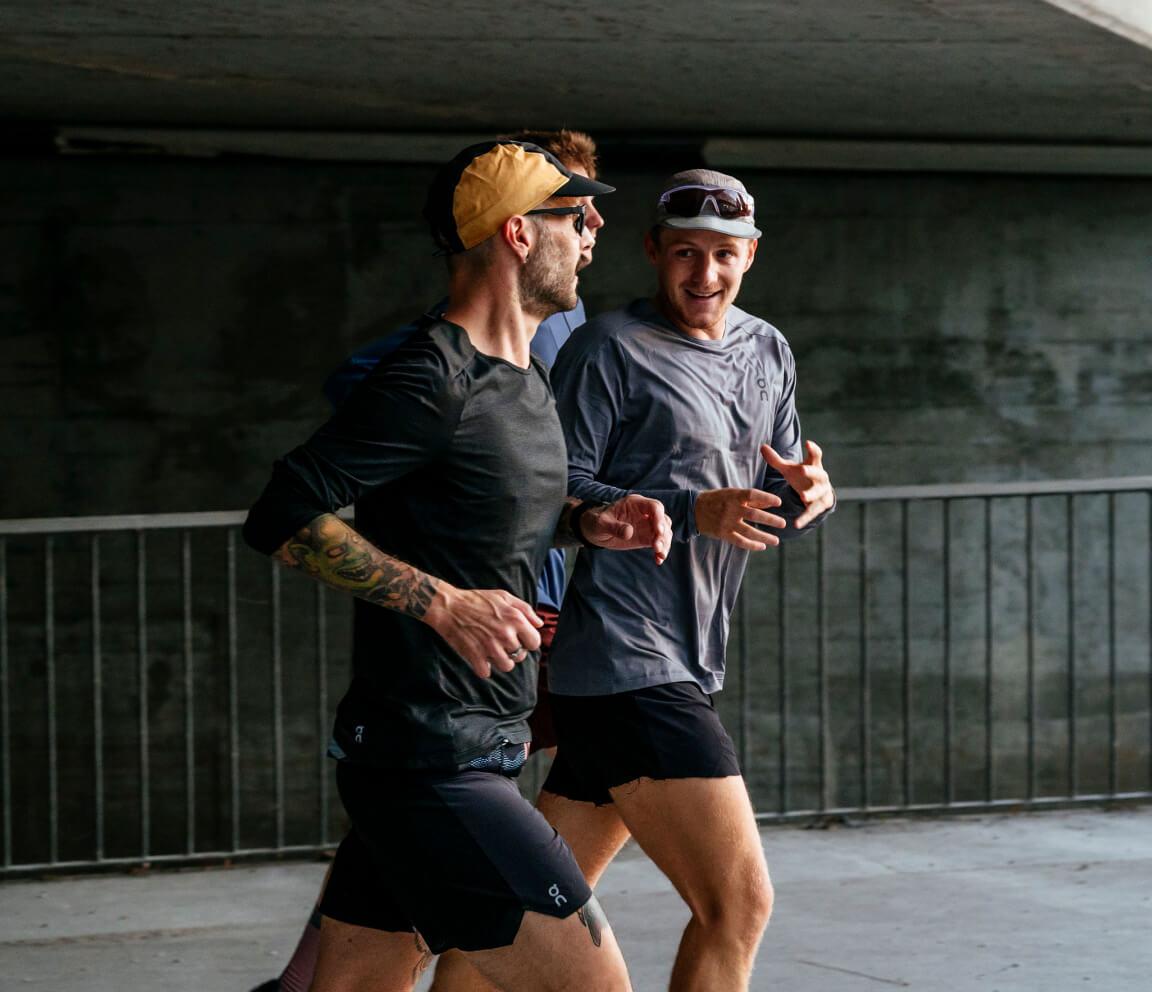Three men in athletic gear jogging together under a concrete overpass, smiling and talking, with a metal railing and dark wall in the background