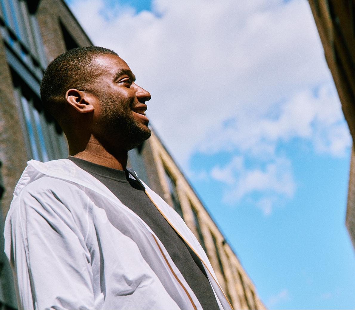 Smiling man in a white jacket stands outdoors between tall buildings under a blue sky.