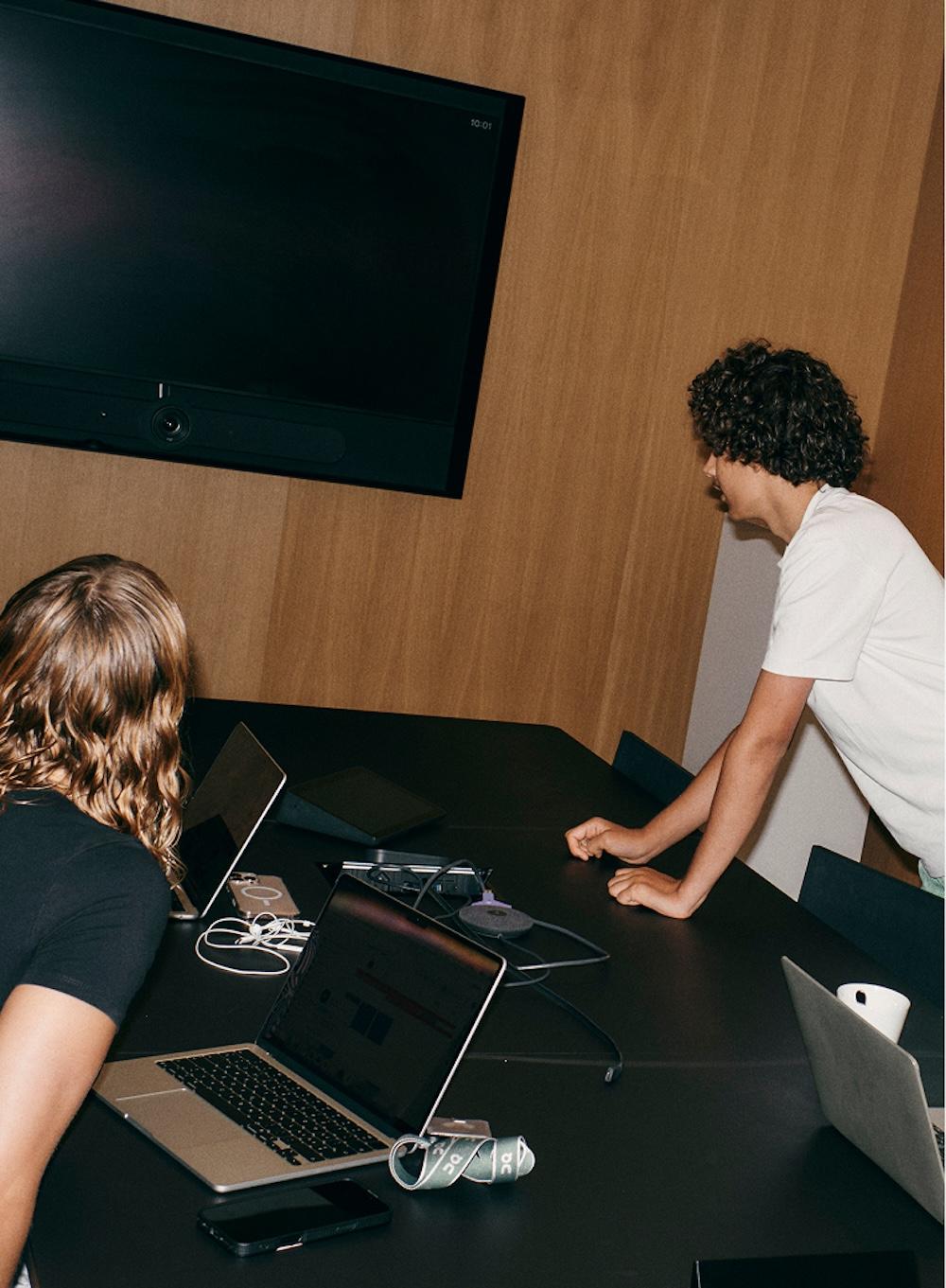 Two people at a conference table with open laptops, cables and a coffee mug, while a large wall-mounted monitor hangs on a wood-paneled wall.
