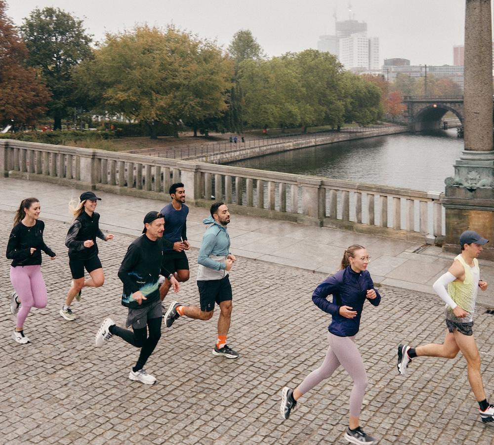 A group of people jogging on a cobblestone riverside promenade with trees and a stone bridge visible in the background.