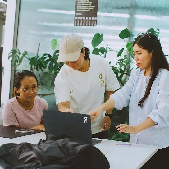 Three people gathered around a laptop at a table—one pointing at the screen, another gesturing, and a third watching—with potted plants visible in the background.