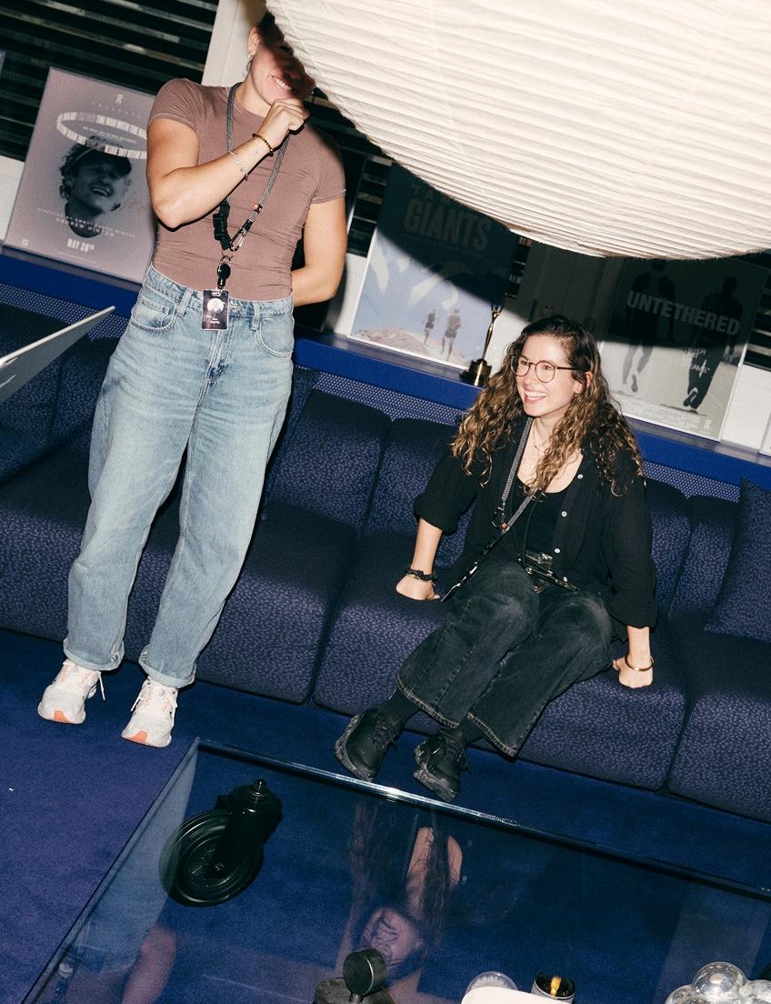 Two women in casual clothes talk and laugh in a lounge with a large hanging light and film posters in the background.