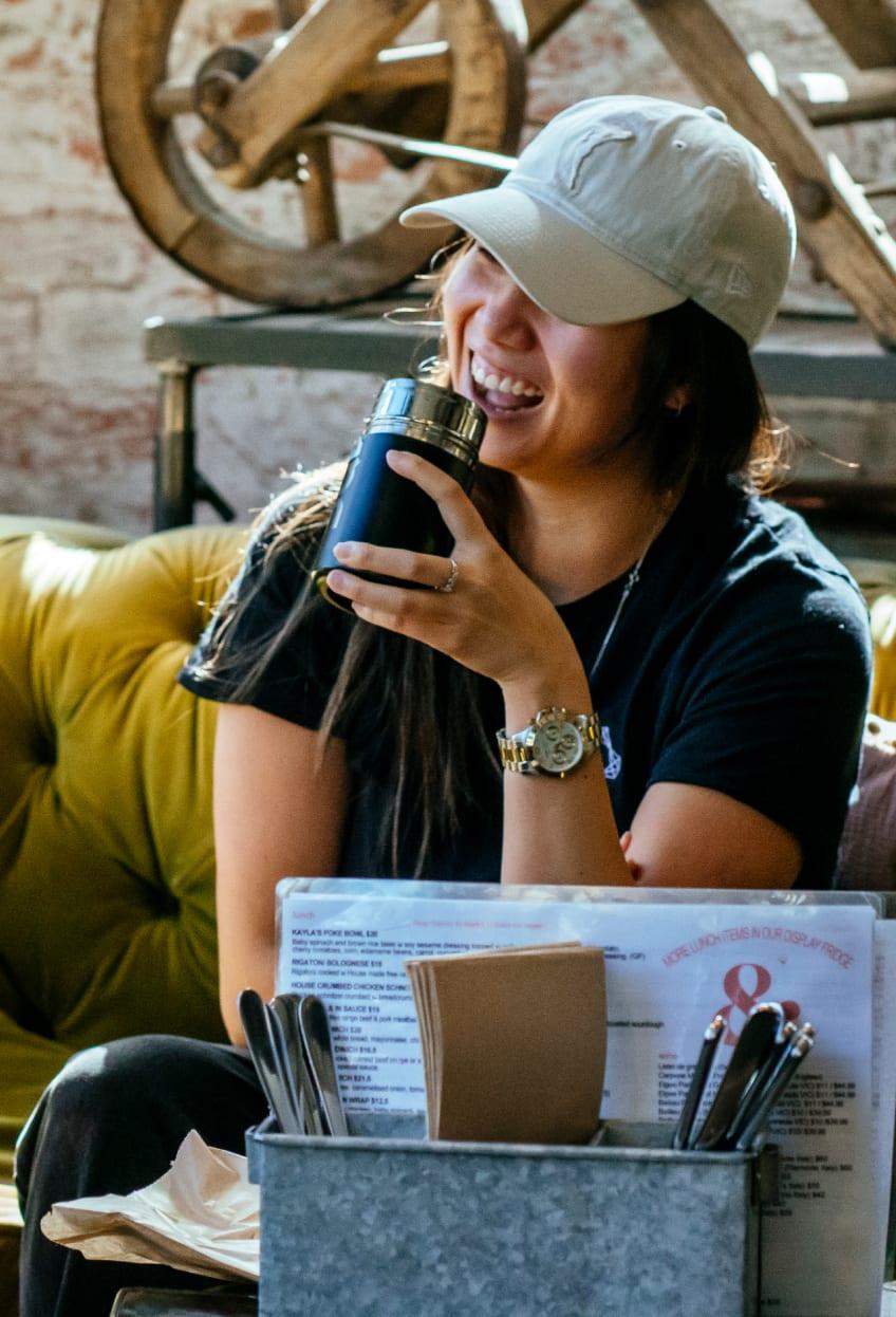 A woman sat on a sofa holding a drinks flask while laughing with someone out of the camera shot.