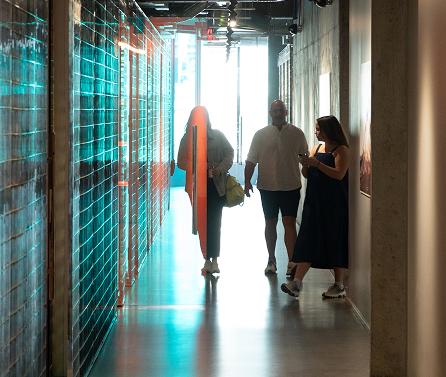 Three people in a narrow modern hallway with a teal-tiled left wall and bright light at the far end, two walking and one leaning against the right wall looking at a phone.
