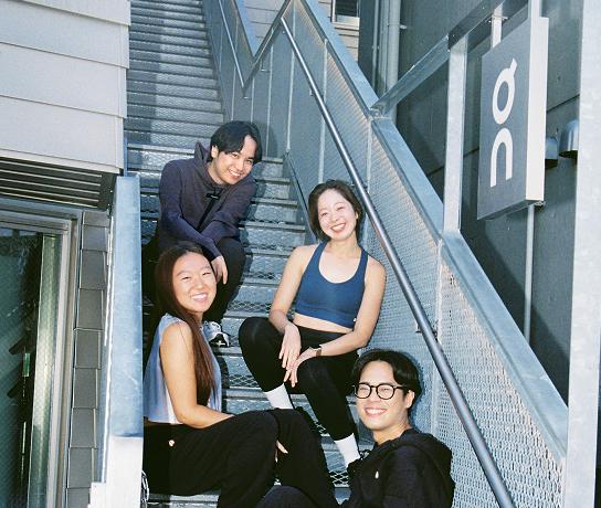 Four smiling people in athletic clothing sit on a metal outdoor staircase beside a railing-mounted sign showing a stylized "on" logo.