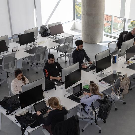 Open-plan office with people working at long communal desks equipped with laptops and large monitors near floor-to-ceiling windows and a concrete support column.