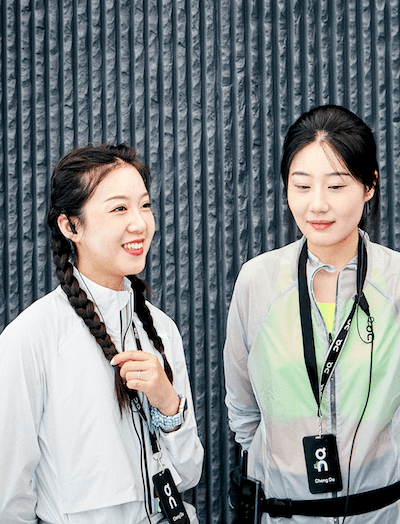 Two women wearing light jackets and lanyards stand in front of a ribbed wall, one smiling with braided hair and the other looking down.