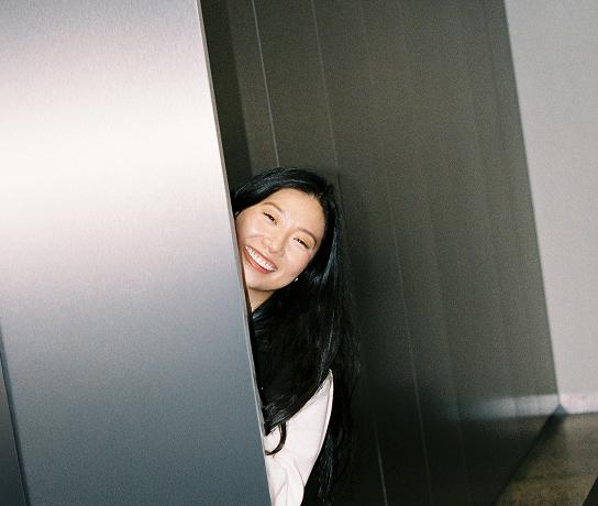 A smiling person with long dark hair peeks from behind a metallic wall panel in a hallway.