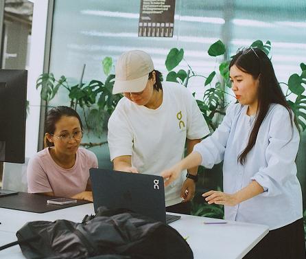 Three people gathered around a laptop at a table—one pointing at the screen, another gesturing, and a third watching—with potted plants visible in the background.