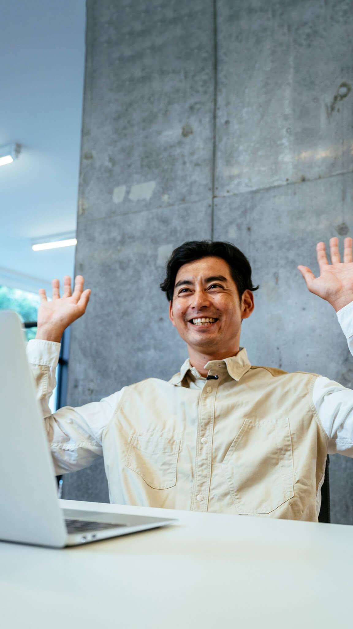 Smiling man sitting at a desk with a laptop, raising both hands in the air, in front of a concrete wall in a brightly lit modern office space.