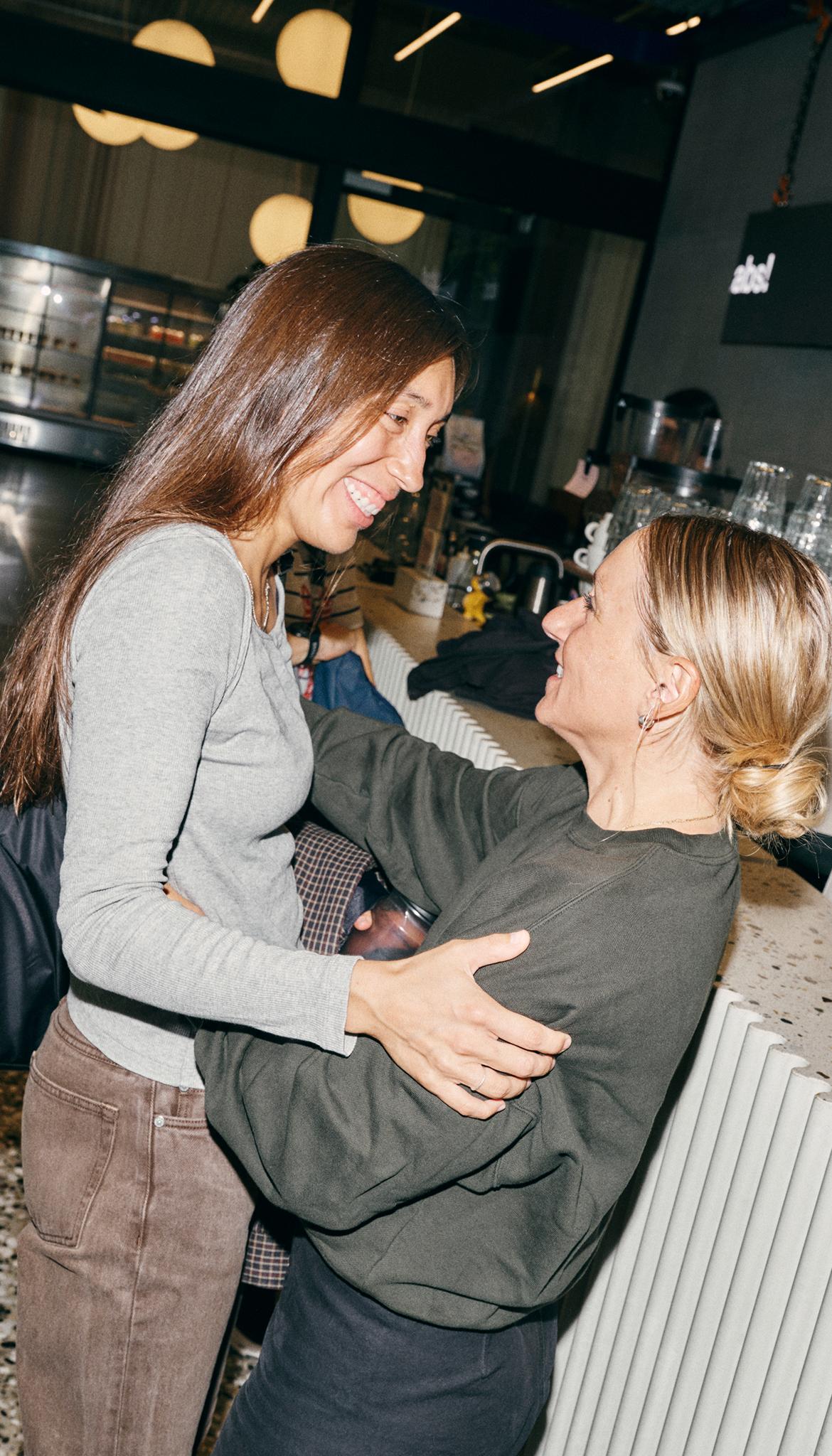 Two women smile and embrace near a café counter, one holding a drink in a glass jar.