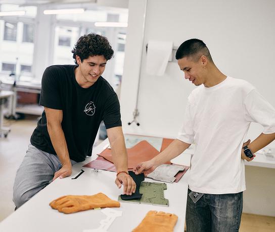 Two men smile while discussing fabric and sole prototypes on a table in a bright workspace, with orange gloves in the foreground.