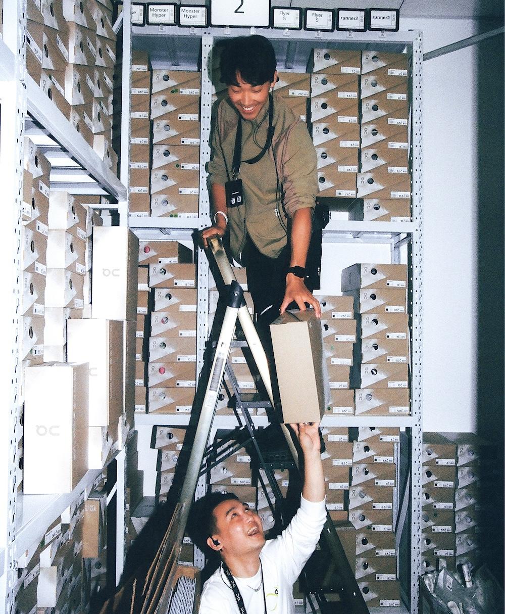 A person standing on a ladder hands a cardboard box to a person reaching up amid metal shelving filled with labeled shoeboxes.
