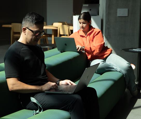 Two people sit on green modular sofas in a dimly lit room, each using a laptop—one wearing a black shirt in the foreground and the other in an orange jacket in the background.