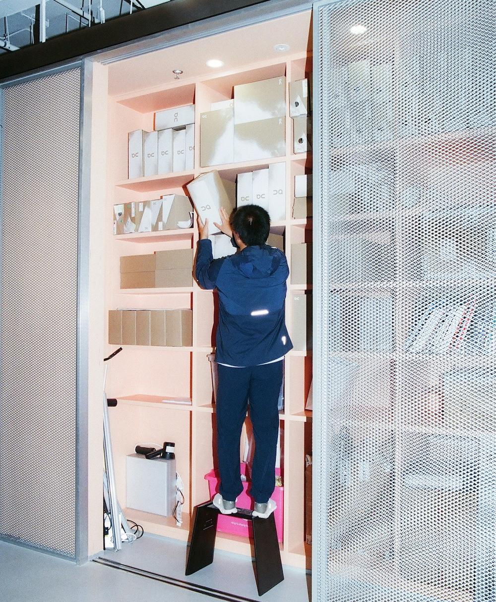 A person standing on two black platforms reaches to place a white box on a pink shelving unit filled with stacked cardboard boxes next to a metal mesh sliding door.