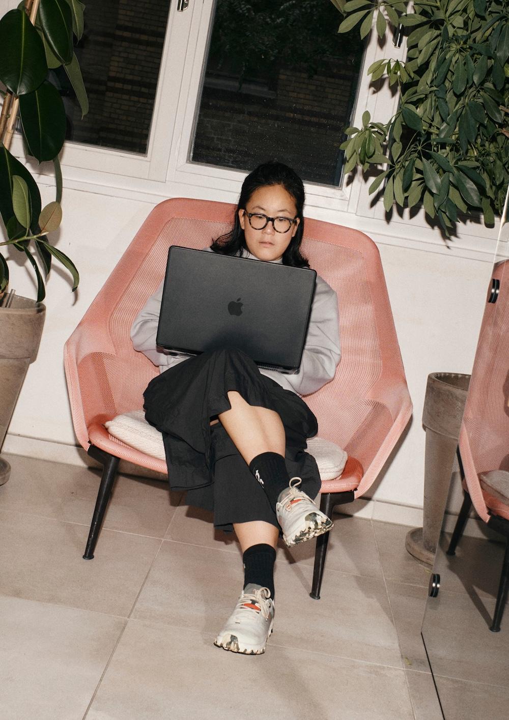 Person sits in a pink chair with legs crossed, focused on a black laptop in a plant-filled indoor space.