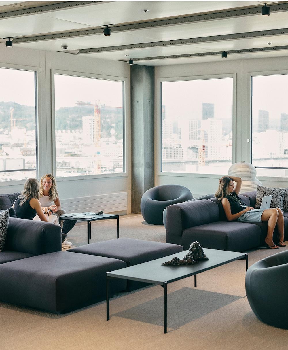 Three women in a modern lounge with large windows and dark modular sofas, two talking at a low table and one reclining on a sofa using a laptop.
