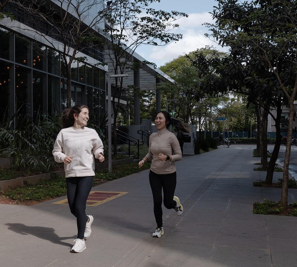 Two women jog and smile on a tree-lined sidewalk beside a modern glass building, wearing light jackets, leggings, and running shoes.