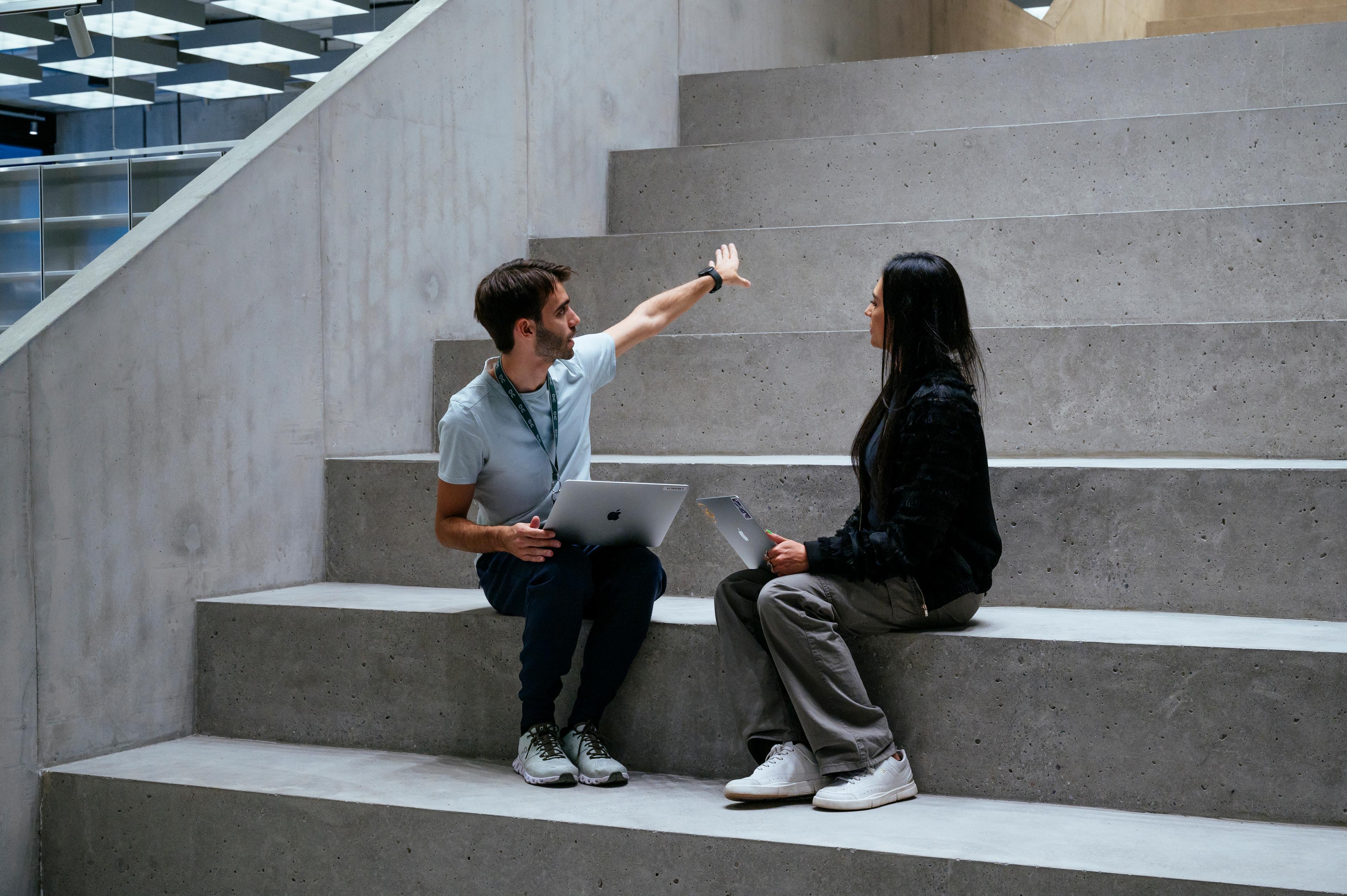 Two people sitting on wide concrete steps indoors, each holding a laptop, as the man gestures upward while speaking to the woman.