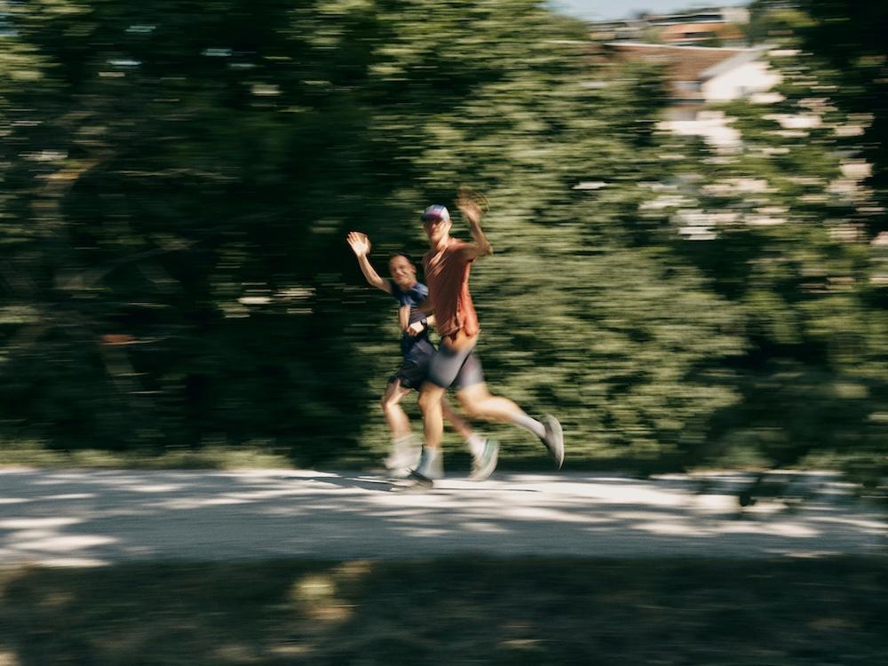 Two runners wave and smile while jogging on a shaded path, captured in motion blur against a backdrop of trees.