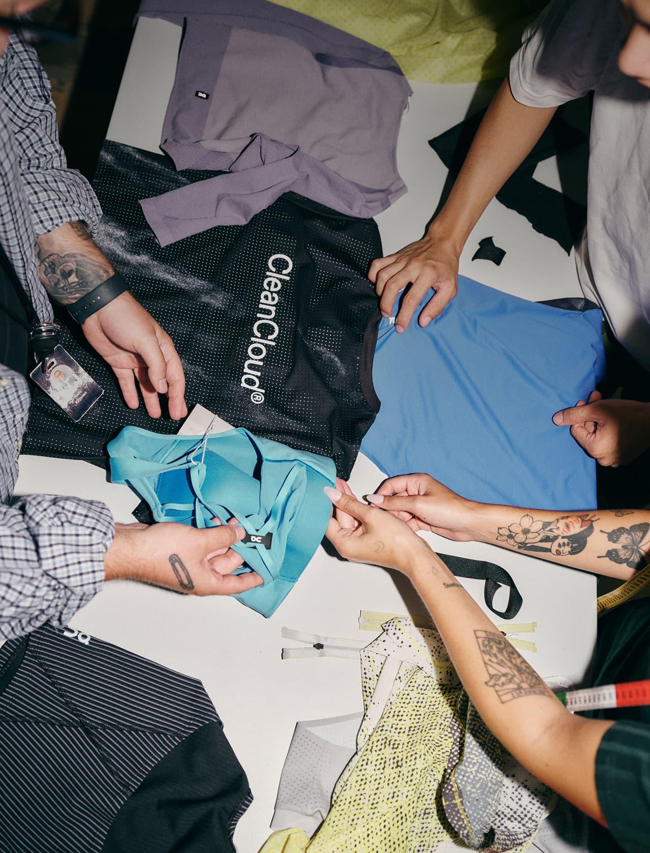 Overhead view of several people’s hands arranging various garments on a white table, including a black mesh piece labeled 'CleanCloud' and blue, teal, and purple clothing.