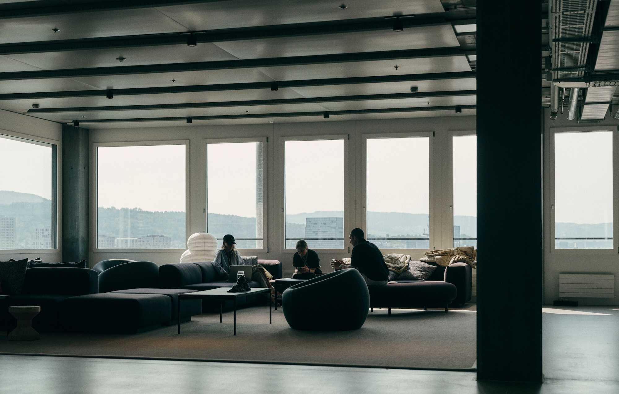Three people sit on low sofas in a spacious, modern lounge with large windows overlooking a distant city and hills.