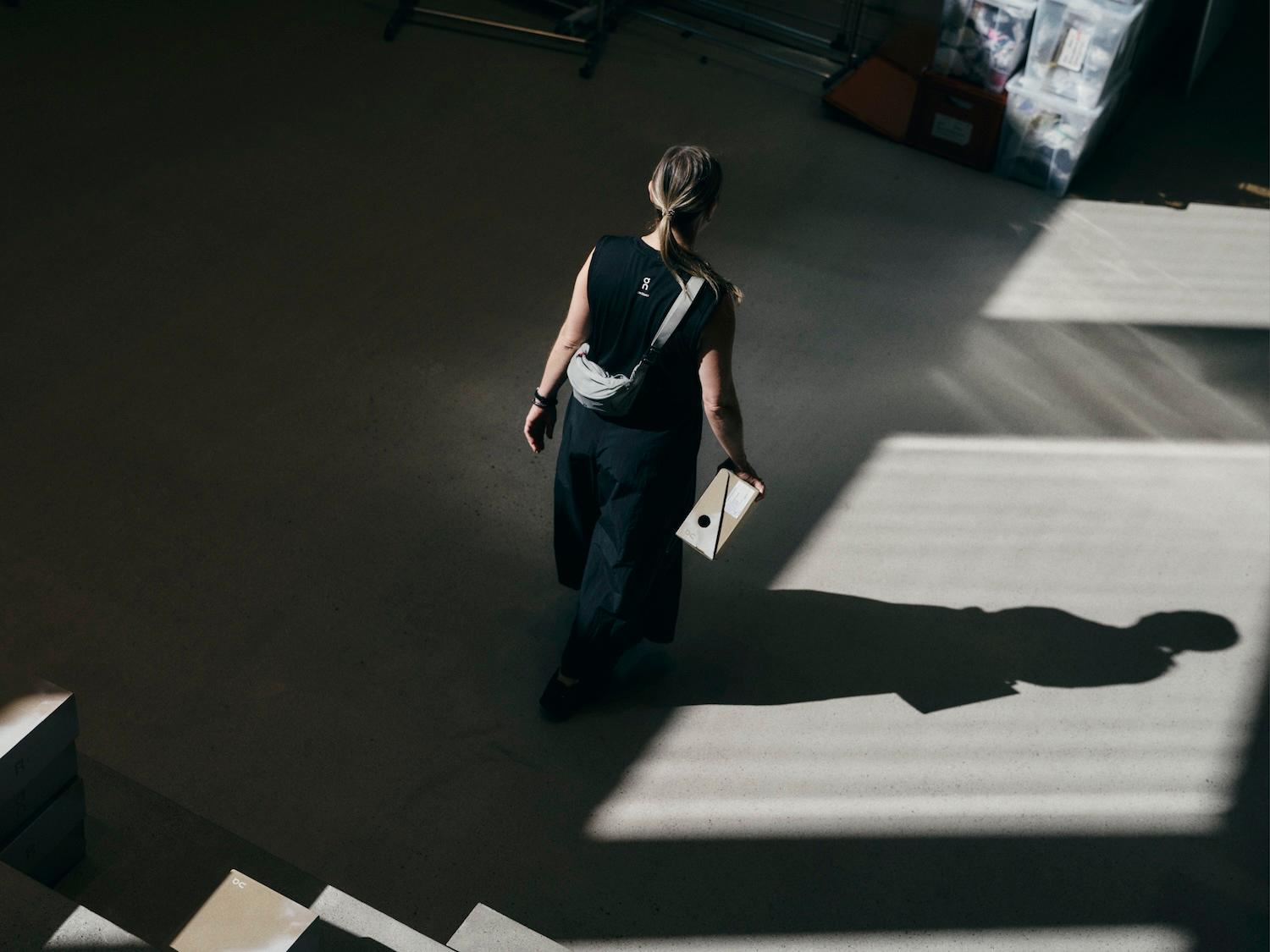 Woman in a black outfit walks across a sunlit room carrying a small cardboard box, casting a strong shadow on the floor.