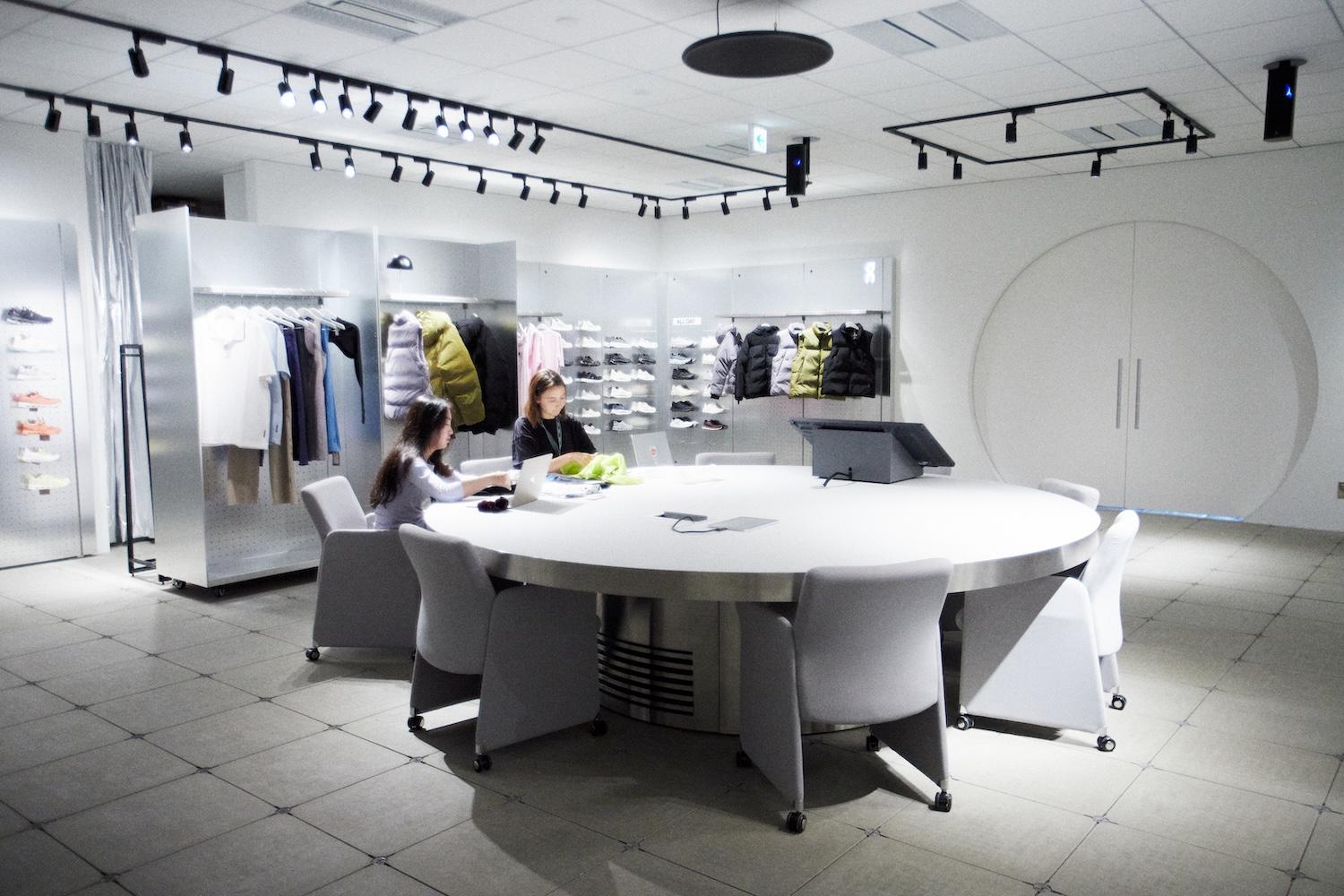 Two women work at laptops around a large round table in a showroom displaying jackets and sneakers on metallic walls.