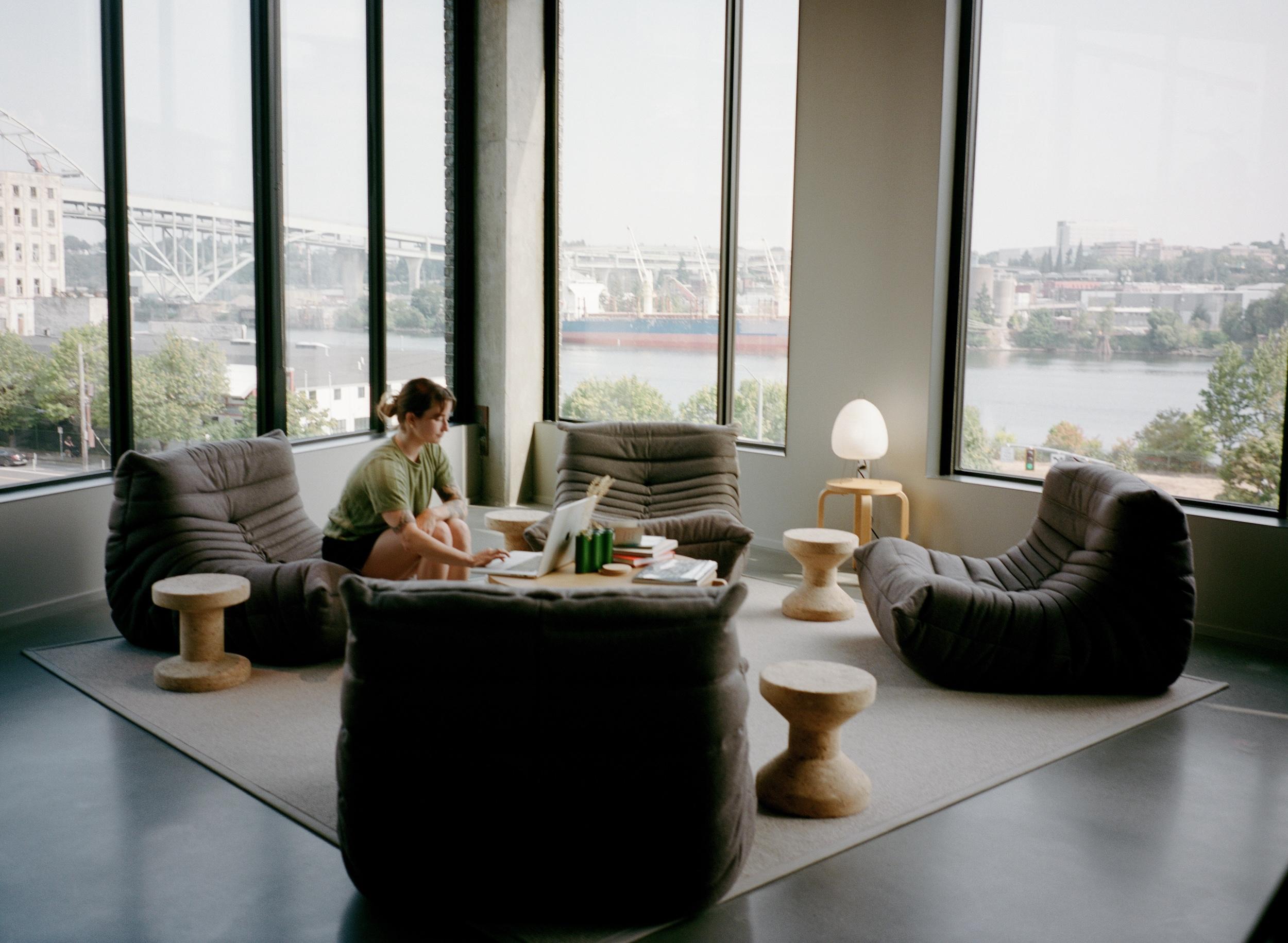 Three people in a narrow modern hallway with a teal-tiled left wall and bright light at the far end, two walking and one leaning against the right wall looking at a phone.