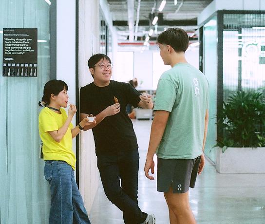 Three people stand and chat in an office corridor: a woman in a yellow shirt eating from a cup, a man in a black shirt leaning against the wall holding a snack, and a man in a green T-shirt and shorts facing them.