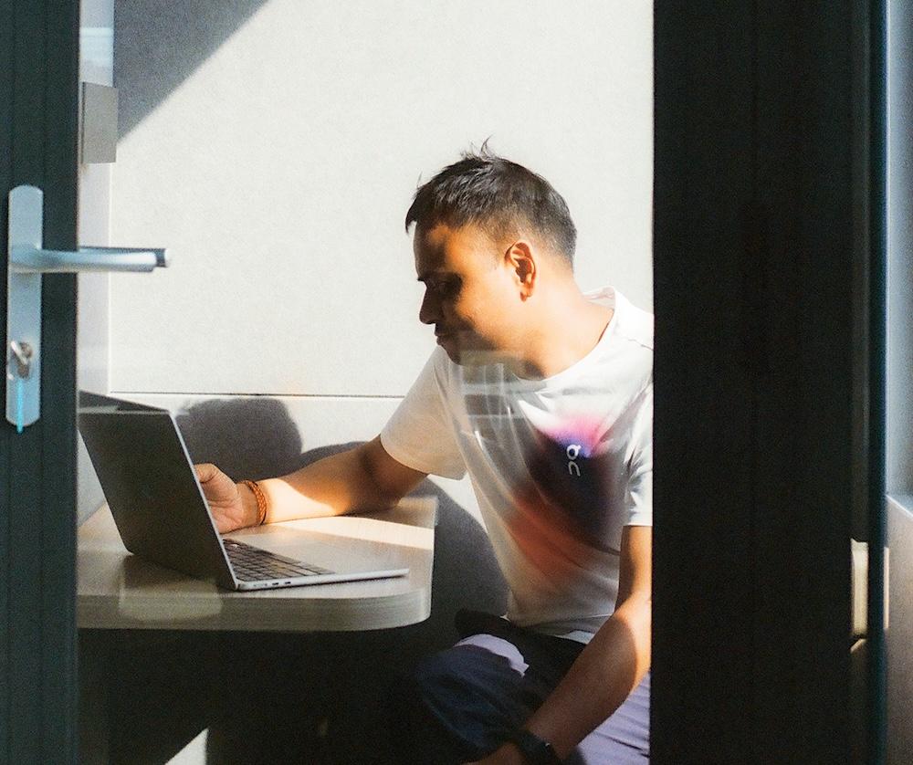 A person sits at a small table using a laptop in a sunlit booth, viewed through a partially open door.