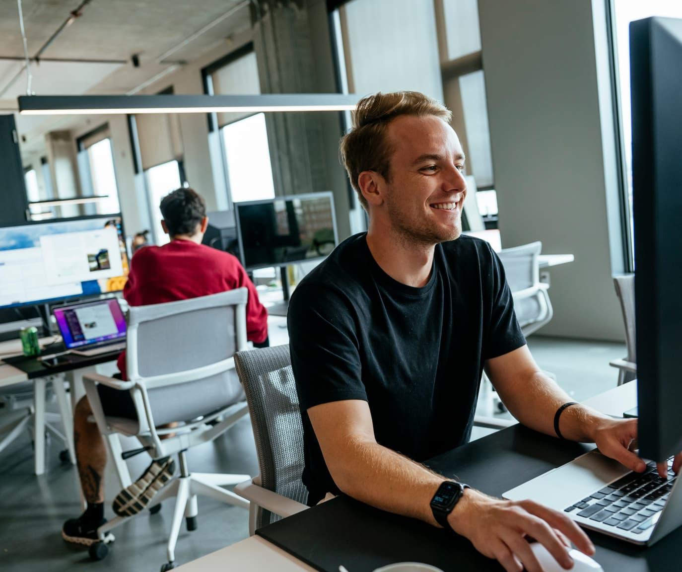 A man at a desk smiling and looking at his computer monitor.