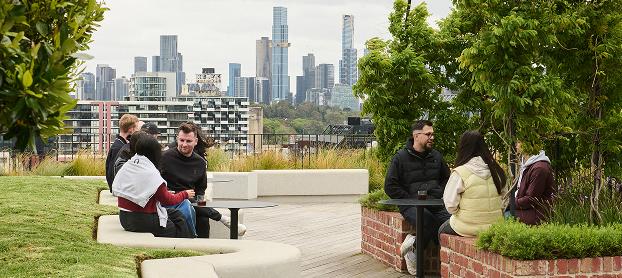 Several people sit and converse at small round tables on a rooftop terrace with planted beds and a city skyline in the background.