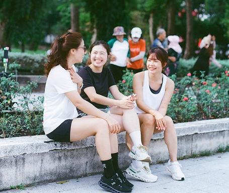 Three women in athletic clothing sit on a low concrete ledge in a park, smiling and talking while people walk in the background.