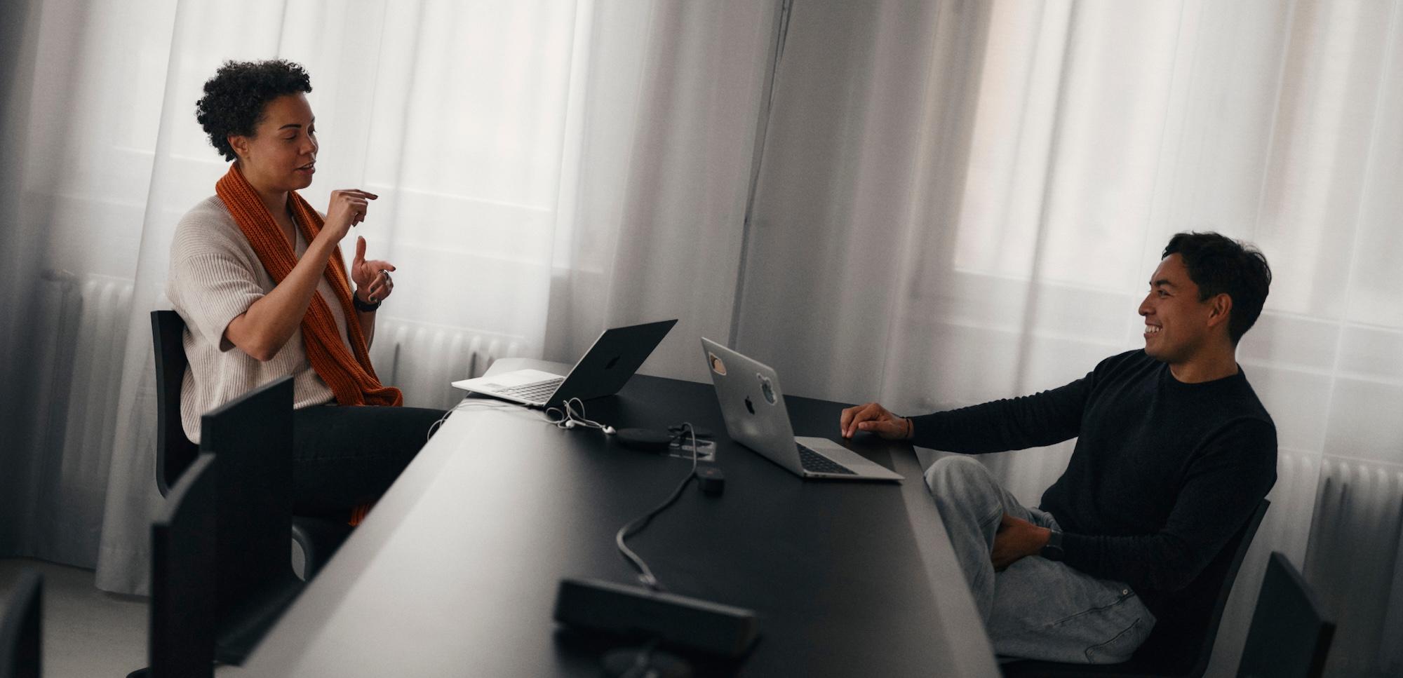 Two colleagues sit at a long table with laptops, having a relaxed conversation in a softly lit room.