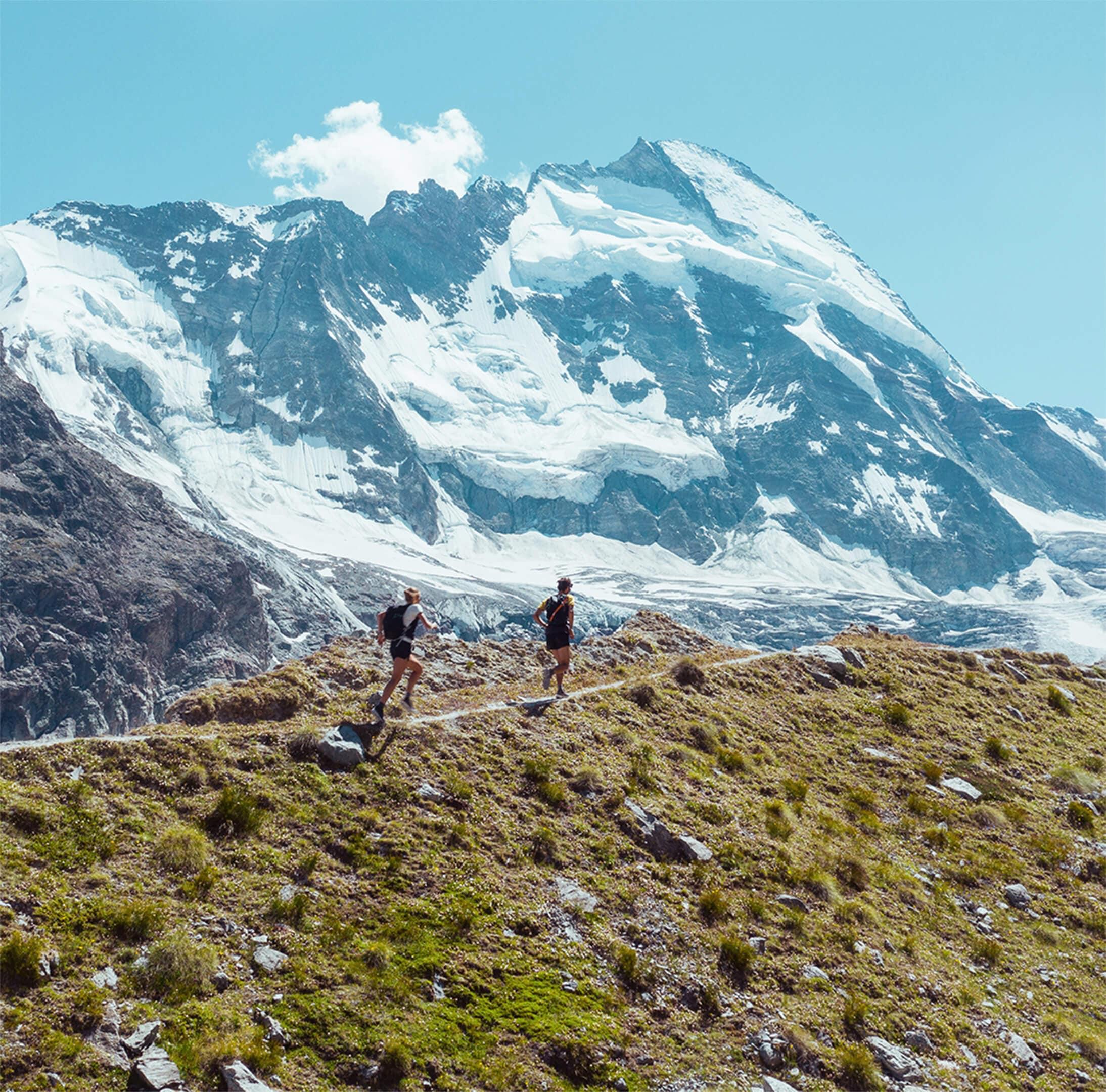 Two hikers walking along a grassy mountain trail with snow-covered peaks towering behind them under a bright blue sky.