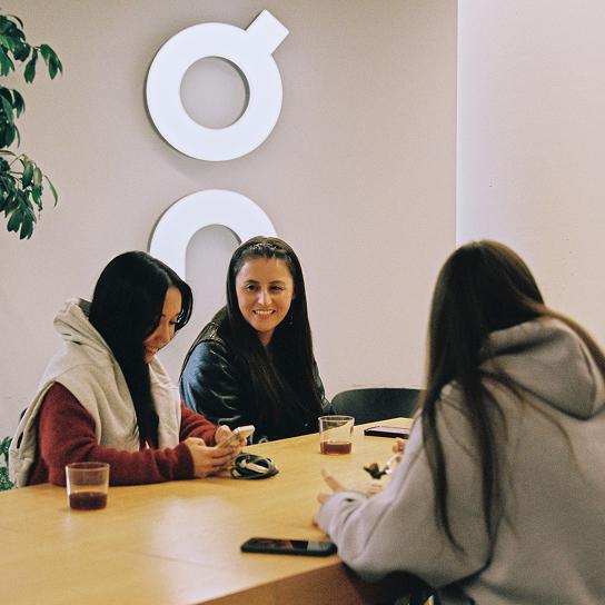 Three people sit around a table with drinks and phones, one smiling while the others look at a phone, with circular white wall shapes visible behind them.