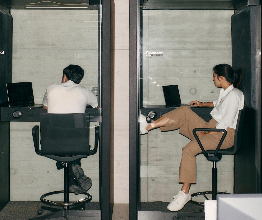 Two people sit in adjacent glass work booths using laptops, one facing away and the other seated with their legs propped on the desk.