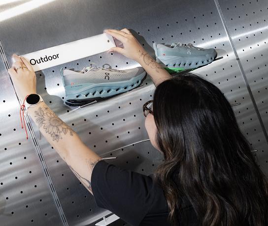 A person with long dark hair and tattooed forearms places a white sign labeled "Outdoor" above two light blue-gray athletic shoes on a perforated metal display wall.