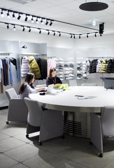 Two women sit at a large round table with laptops in a brightly lit clothing and shoe showroom where jackets and sneakers are displayed on wall-mounted shelves.