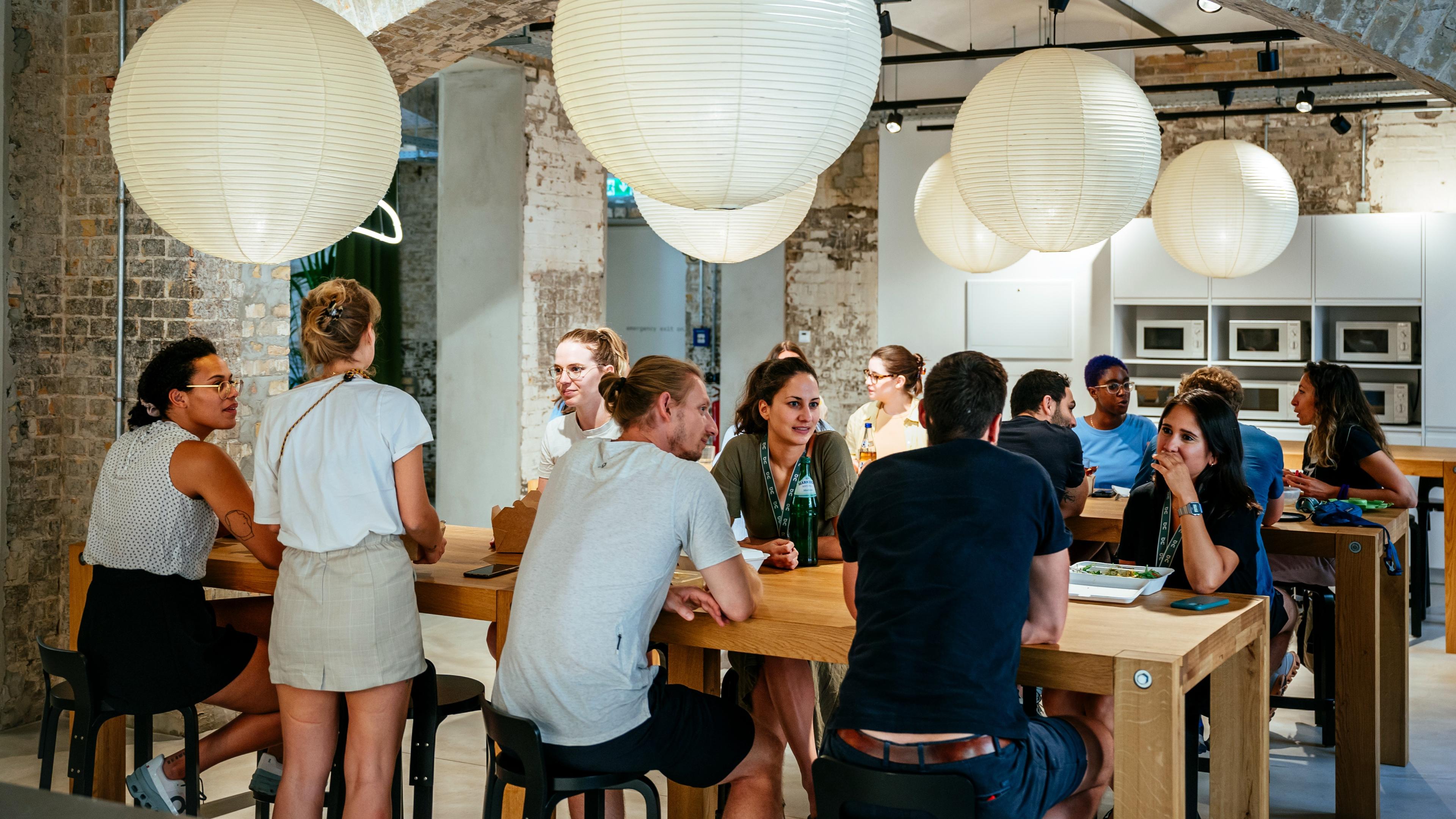 Groups of people sit and chat around wooden tables in a brick-walled communal space with hanging paper lanterns and a wall of microwaves in the background.