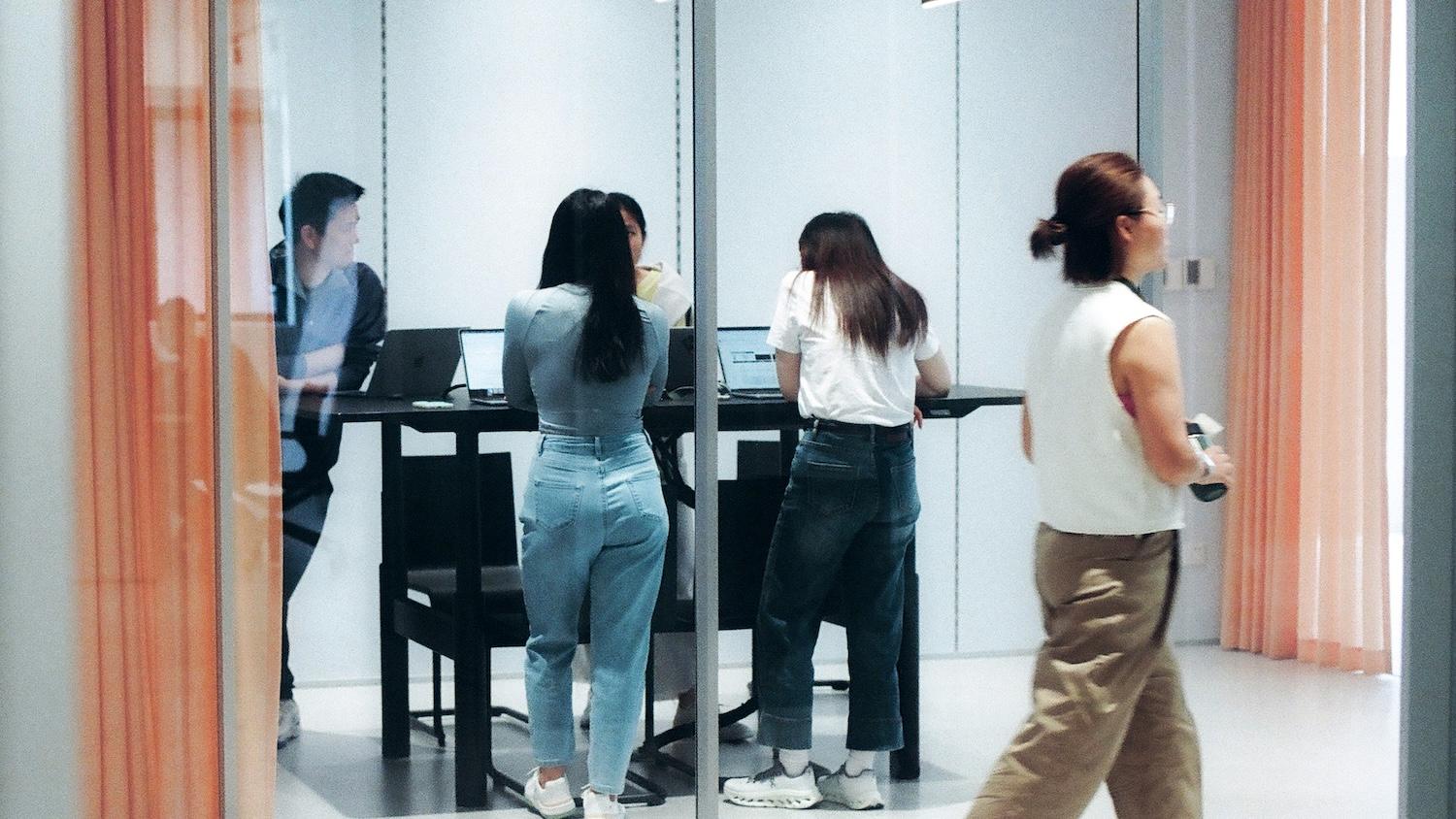 Four people work on laptops around a high table in a glass meeting room, while another person walks by outside holding a drink.
