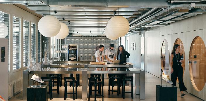 Open-plan modern workspace with a long stainless-steel table and stools where three people stand looking at a laptop beneath large round pendant lights, with shoe displays on pedestals and circular wooden doors along the right wall.