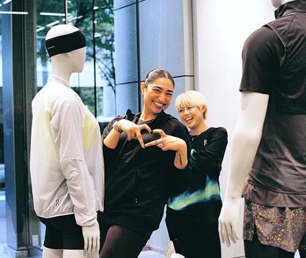 Two smiling people stand between two mannequins in a clothing store window display and make a heart shape with their hands.