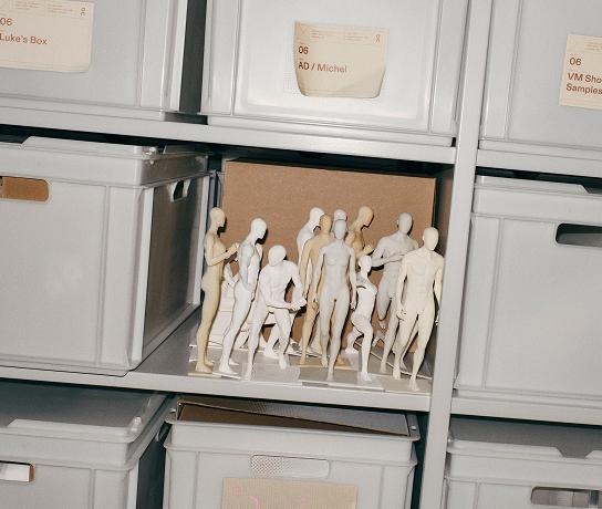 A shelving unit of white storage bins with a group of small unpainted human-shaped mannequins standing together on an open shelf in front of a cardboard box.