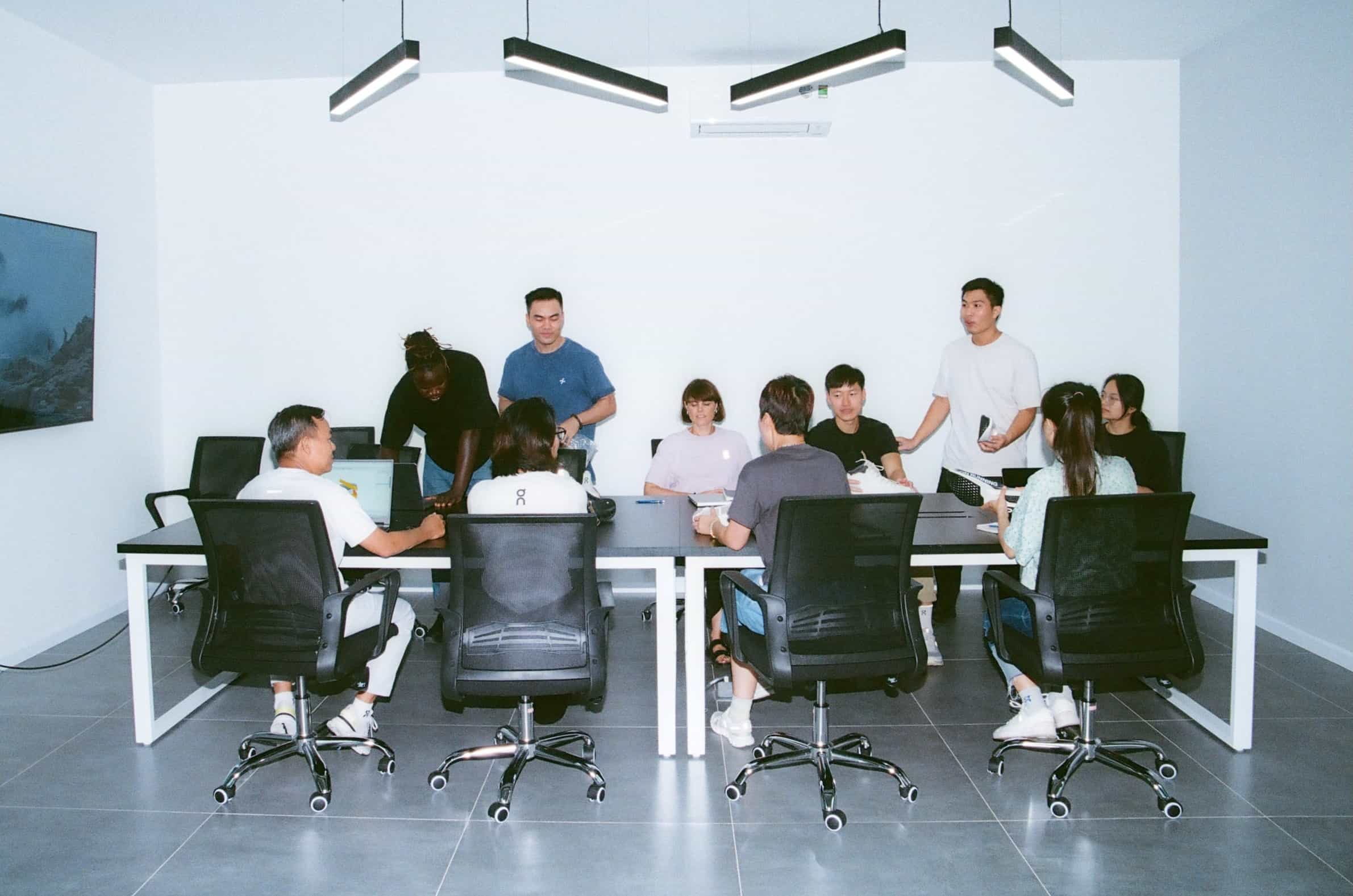 A diverse group of people seated and standing around a conference table in a bright meeting room.