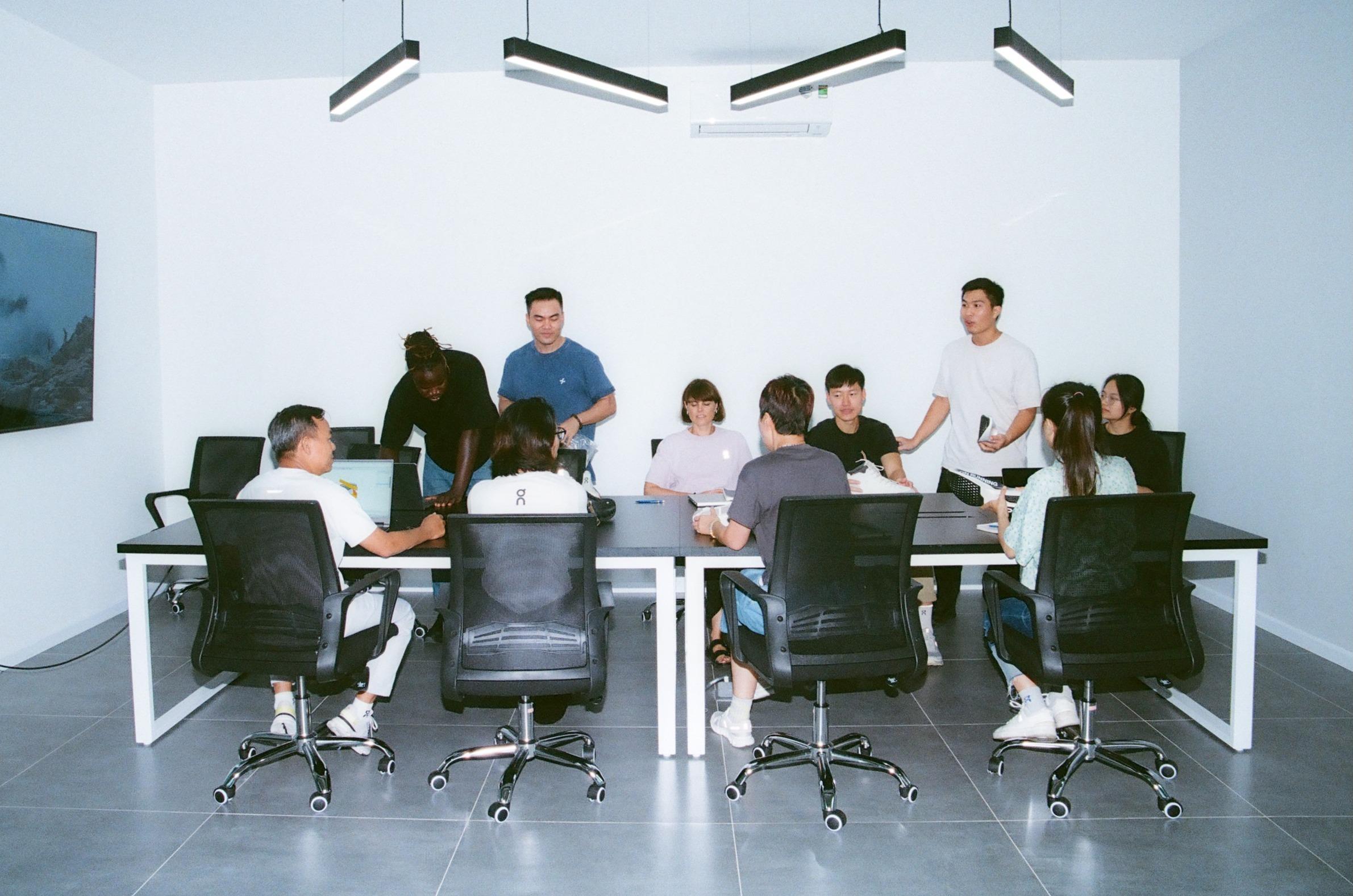 A diverse group of people seated and standing around a conference table in a bright meeting room.